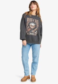 Felpa color carbone con una stampa grafica che presenta elementi solari e floreali; abbinata a jeans azzurri chiari e scarpe slip-on marroni.