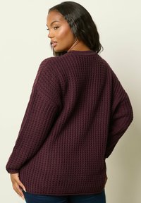 Frau mit dunklen Haaren, die einen strukturierten, übergroßen dunkelvioletten Pullover trägt, posiert mit dem Rücken zugewandt und dem Kopf leicht zur Seite gedreht.