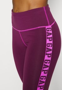 Vinröda leggings med hög midja, som har en framträdande rosa logotryck längs sidan och kontrasterande rosa sömmar.
