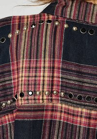 Tessuto a quadri con righe rosse, rosa e beige presenta borchie tonde argentate e nere disposte in file orizzontali su uno sfondo scuro.