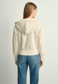 Krótkie kremowe bluza z kapturem, z prążkowanymi mankietami i dołem, w połączeniu z jasnoniebieskimi dżinsami. Gładka faktura materiału i subtelne detale przeszyć.