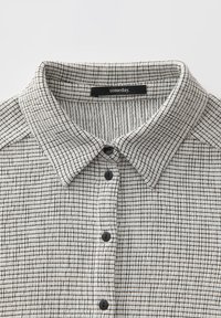 Camicetta a maniche corte con colletto in un tessuto bianco testurizzato con motivo a griglia nera, dotata di cinque bottoni neri e un'etichetta con la scritta "un giorno".