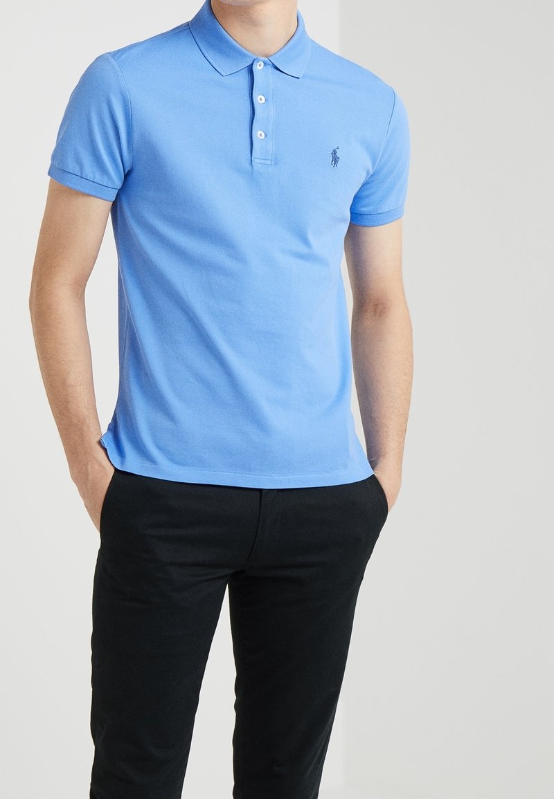 Homme portant un polo bleu clair à manches courtes avec un petit logo sombre sur la poitrine et un pantalon noir, mains dans les poches, fond clair.
