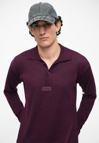 Figūrainis burgundijas garām piedurknēm polo krekls ar apkakli un trim pogām, ar zīmola uzlīmi. Modelis valkā pelēku cepuri ar izliekto malu.