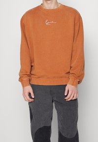 Persona con un sudadera de manga larga de color naranja desgastada con un logo en blanco y pantalones gris oscuro con parches negros, de pie contra un fondo liso.