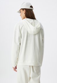 Felpa beige chiaro con cappuccio, realizzata in tessuto morbido. Presenta una vestibilità rilassata e polsini a costine, abbinata a un cappellino coordinato.