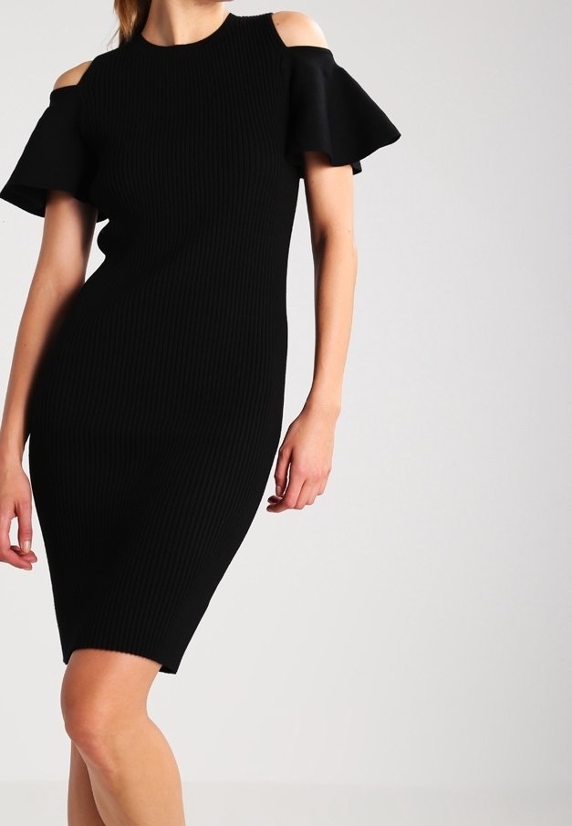 Femme portant une robe côtelée noire longueur genou avec découpes aux épaules dénudées et manches courtes évasées sur fond uni.