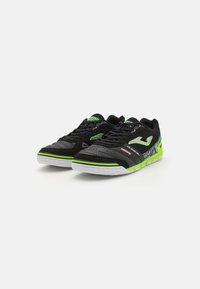 Des chaussures de sport noires avec des accents verts, présentant une tige texturée, des sections perforées et une semelle en caoutchouc blanche. Le design comprend un détail de marque.