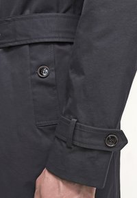 Tmavě námořnický trenčkot s hladkou texturou, dvojitým knoflíkovým zapínáním a nastavitelnými cuff strapami s knoflíky.