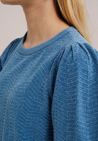 Suéter azul texturizado con un patrón de espiga, cuello de canalé y mangas abullonadas. La tela parece suave y ligera.