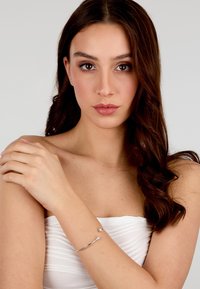 Bracciale d'argento con due pietre trasparenti, design curvo, texture liscia, indossato al polso di una modella con lunghi capelli scuri. Top bianco senza spalline.