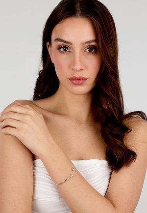 Bracciale d'argento con due pietre trasparenti, design curvo, texture liscia, indossato al polso di una modella con lunghi capelli scuri. Top bianco senza spalline.