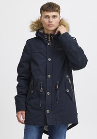 Marineblå parka-jakke med faux fur hætte, frontknapper, snøre i taljen og flere lommer. Holdbart stof med en glat tekstur.