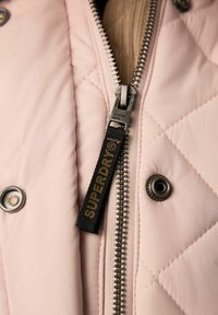 Różowa kurtka pikowana z srebnym zamkiem błyskawicznym, posiadająca czarny uchwyt do zamka z napisem "SUPERDRY" oraz detal z zatrzaskiem.