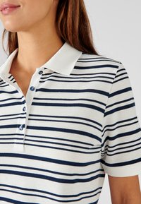 Camisa polo a rayas en blanco y azul marino, con cuello tipo polo, botonera y mangas cortas, realizada en una tela suave y texturizada.