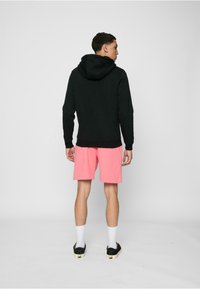 Svart hettegenser med kengurulomme, båret over rosa shorts, sett bakfra. Har en løs passform og ribbestrikkede mansjetter.