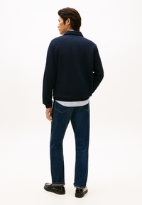 Veste sweat bleu marine avec col, poignets et ourlet côtelés, associée à un jean denim bleu foncé et des chaussures noires. Fond uni.