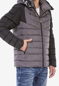Chaqueta acolchada gris y negra con frontal con cremallera, texturas acanaladas y capucha; presenta paneles en ángulo y bolsillos laterales.