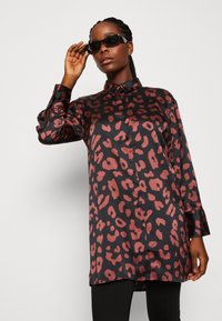 Juodas sateninis marškinis su raudonu leopardų raštu, paspaudžiamas priekyje, ir ilgomis rankovėmis. Modelis nešioja saulės akinius, stiliaus derinys su juodais legingais.