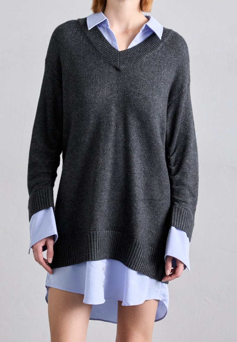 Femme portant un pull ample gris foncé à col en V superposé sur une robe chemise bleu clair oversize sur un fond uni.