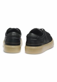 Black leather sneakers with a textured design, low heel, and tan rubber sole. Features branding on the heel and side detailing.