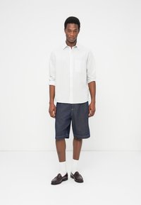 Λευκό ριγέ πουκάμισο με μακριά μανίκια, μπλε τζιν σορτς, λευκές κάλτσες και σκούρα μοκασίνια. Το μοντέλο στέκεται σε απλό φόντο.