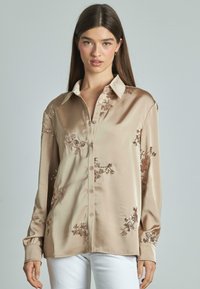 Camicia beige in satin con maniche lunghe, chiusura frontale con bottoni, ricamo di paillettes floreali e colletto classico. Abbinata a pantaloni bianchi.