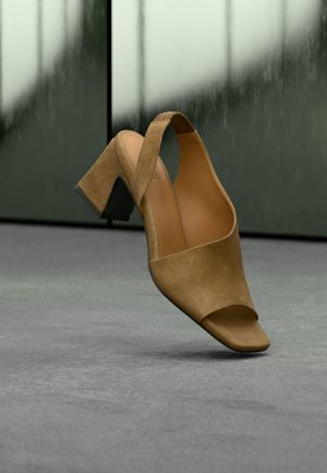 Tan semsket åpen tå slingback blokkhæl sko hengende over en grå overflate med uklare grønne paneler i bakgrunnen.