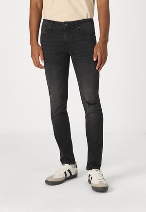 Hombre vistiendo jeans negros ajustados con un pequeño rasgón en la rodilla derecha, camisa beige y zapatillas blancas con rayas negras sobre un fondo blanco.