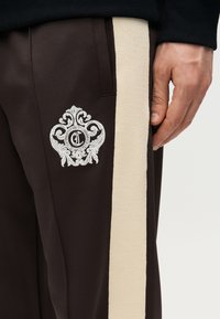 Δεξί χέρι δίπλα σε σκούρο καφέ παντελόνι με φαρδιά μπεζ πλαϊνή ρίγα και λευκό κεντημένο περίτεχνο έμβλημα κοντά στην εμπρόσθια τσέπη.