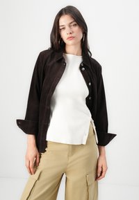 Brown corduroy button-up jacket, white ribbed top, and light beige cargo pants. Features rolled sleeves and side buttons on the top.