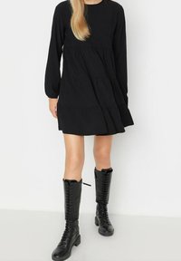 Abito lungo nero a maniche lunghe con design a strati, tessuto testurizzato e vestibilità rilassata, abbinato a stivali neri al ginocchio con lacci.