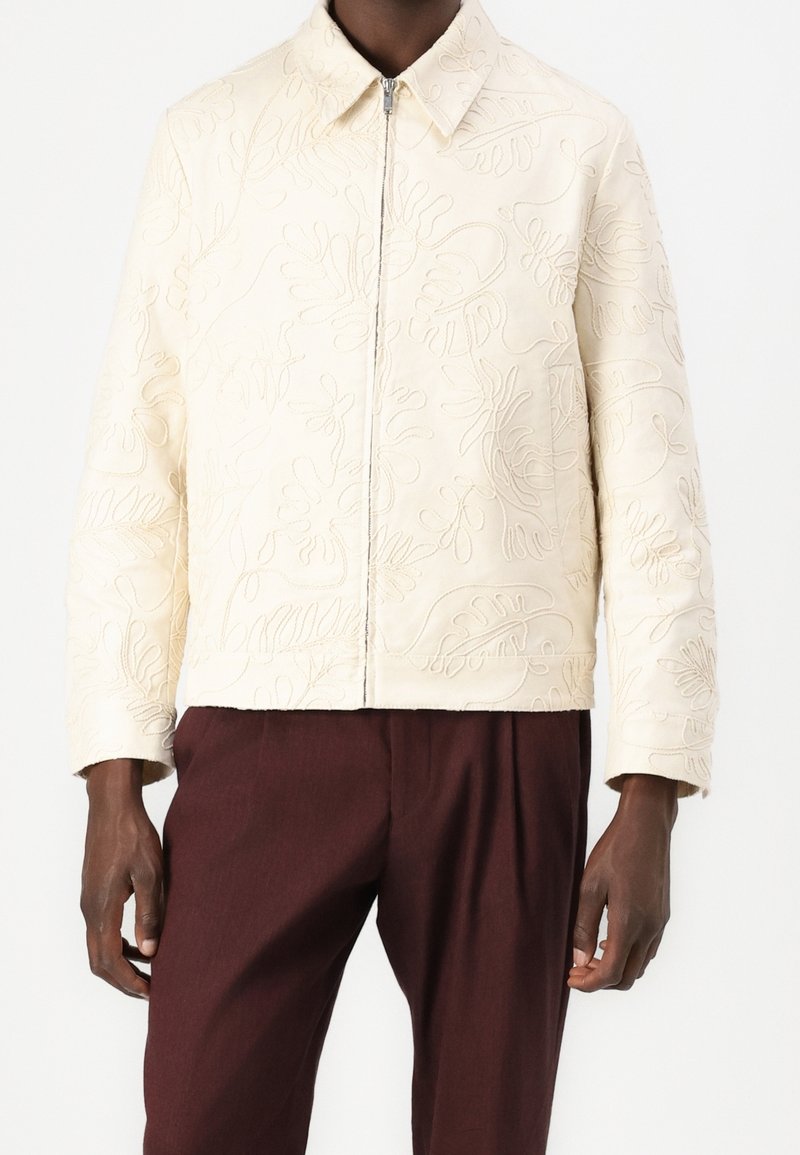 Krämfärgad kviltad jacka med blommönsterbroderi, med dragkedja och spetsig krage. Matchas med mörkbruna byxor.