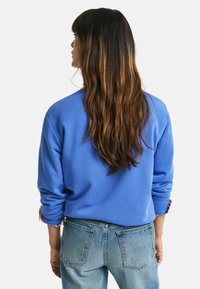 Felpa blu con polsini a costina, abbinata a jeans chiari. La felpa presenta una vestibilità rilassata e una texture morbida. Vista posteriore.