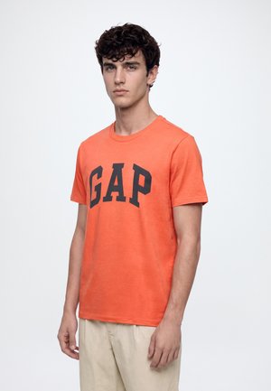 Jeune homme aux cheveux bouclés et foncés, portant un t-shirt orange GAP et un pantalon beige, debout devant un fond clair uni.