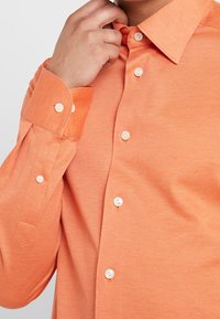 Camisa de manga larga naranja con un tejido texturizado, que presenta un cuello abotonado, botones blancos y un puño ajustable con detalles.