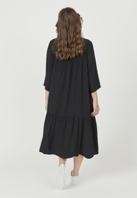 Svart klänning med trekvartslång ärm och en lager-på-lager nederdel. Slät tyg, lös passform, bärs med vita sneakers. Bakifrån syns detaljer med veck.