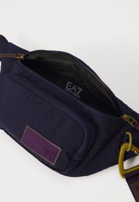Jūrinio nailono fanny pack'as su tekstūruotu paviršiumi, turintis priekinį užtrauktuką, violetines detales ir geltoną metalinį sagtį. Viduje yra prekės ženklo etiketė.