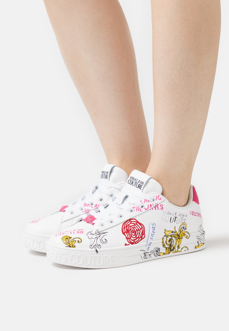 Sneaker bianchi con design grafici colorati, inclusi accenti rosa, motivi floreali e artistici, e una suola testurizzata etichettata "VERSACE JEANS COUTURE."