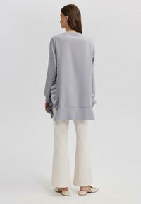 Felpa grigia chiaro con dettagli arricciati sui lati, maniche lunghe e orlo curvo, abbinata a pantaloni flared color crema e scarpe neutre.