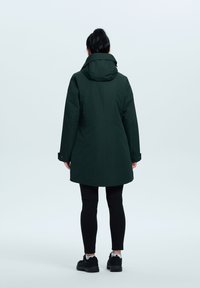 Chaqueta impermeable verde oscuro con capucha, con un corte recto y dos bolsillos laterales, combinada con leggins negros y zapatillas deportivas.