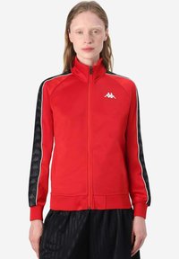 Giacca tuta rossa con zip, con dettagli a strisce nere e bianche e logo Kappa sul petto; tessuto morbido con design aderente.