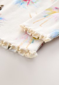 Geribbelde witte stof met zachte pastel aquarelpatronen. De mouwen hebben geplooide manchetten aan de uiteinden, wat het speelse ontwerp versterkt.