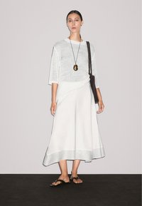 Model staat in een lichte, luchtige outfit met een losgebreid topje en een vloeiende rok, accessorized met een lange ketting en een gevlochten tas.