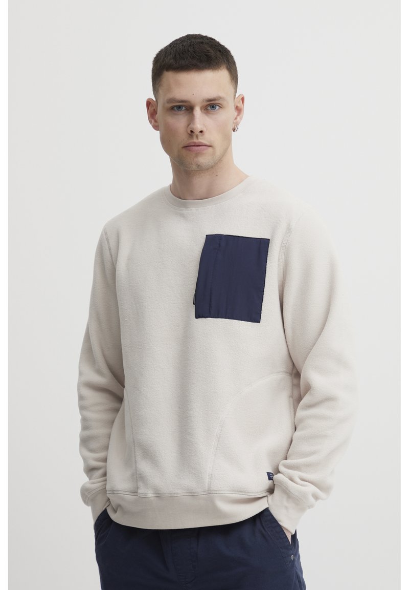 Sudadera beige claro con un bolsillo azul marino. Confeccionada con una textura suave, escote redondo y bolsillos laterales. Corte casual con puños acanalados.