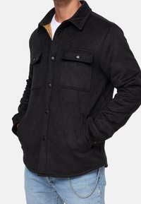 Czarna kurtka koszulowa z wełny z ostro zakończonym kołnierzem, dwoma przednimi kieszeniami, zatrzaskami i beżową podszewką, noszona na białej koszuli.