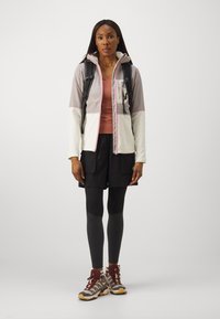 Ελαφρύ, πολύχρωμο μπουφάν με φερμουάρ, κουκούλα και τσέπες. Συνδυάζεται με καφέ μπλούζα, μαύρες βερμούδες και σκούρες κολάν, παρέχοντας υποδήματα πεζοπορίας.