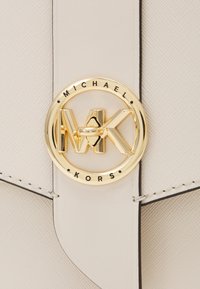Primo piano della chiusura con il logo oro di Michael Kors su una borsa in pelle beige chiaro dalla texture particolare.