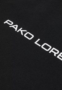 Czarna tkanina o gładkiej fakturze, na której wyraźnie widnieje biały napis "PAKO LORET" w wyboldowanej, nowoczesnej czcionce.