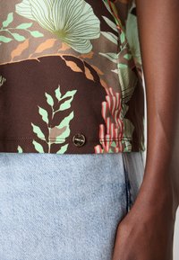 Kortermet brun blomstret topp med grønne og røde detaljer; har et dekorativt metalllogo og et glatt tekstilmateriale. Kombinert med lyse jeans.